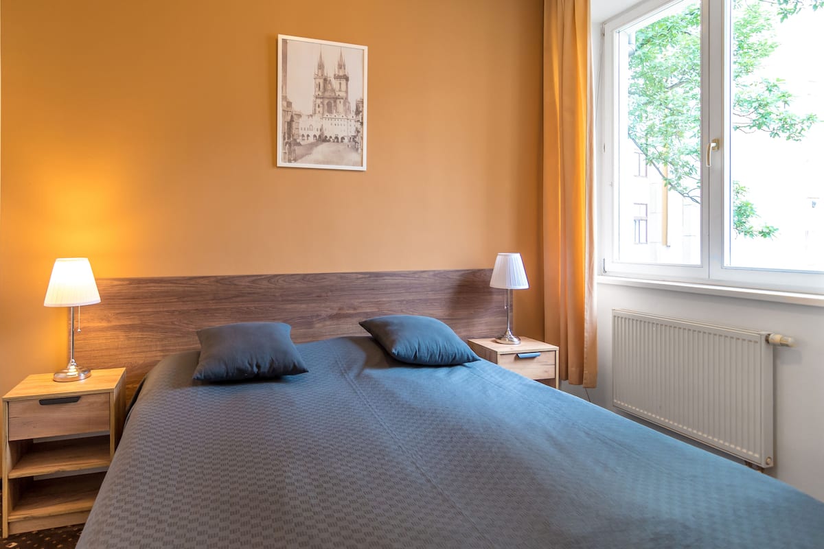 A neatly made bed with a headboard, two bedside lamps, a nightstand, and a window with curtains.