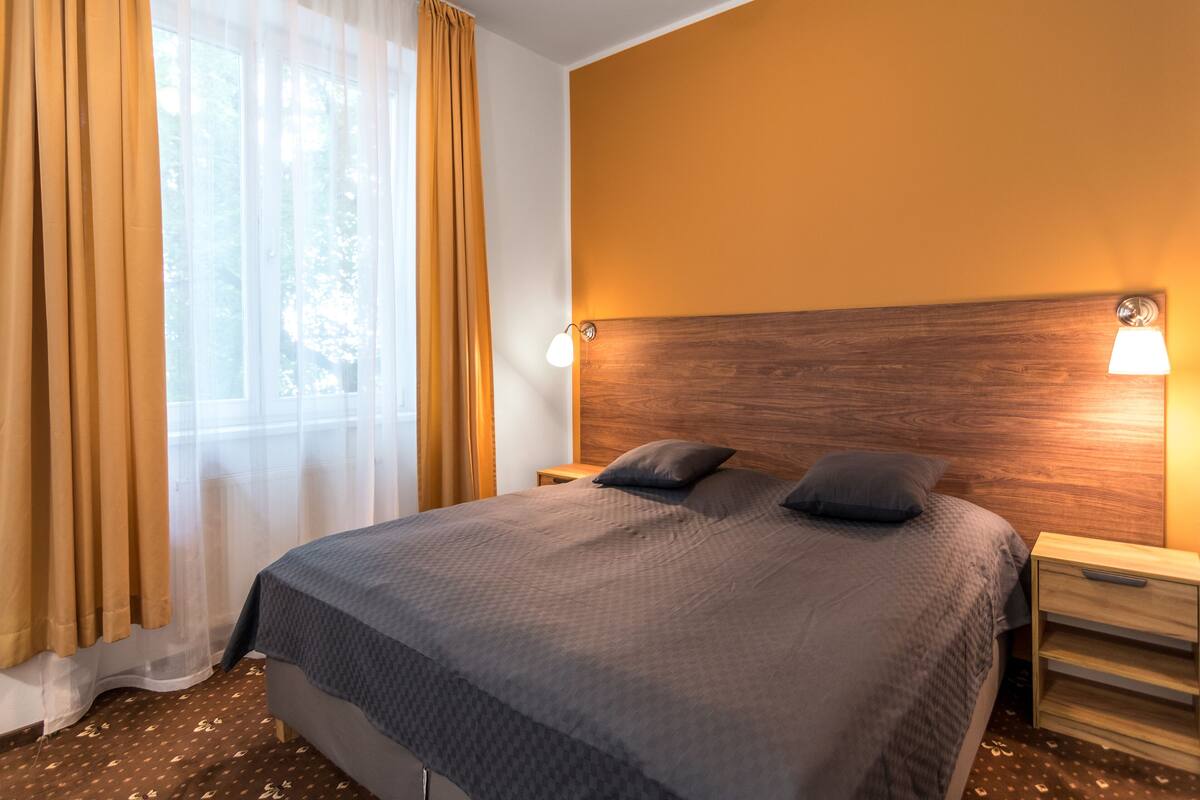 A hotel room with a large bed, wooden headboard, two bedside tables, and a window with sheer curtains.