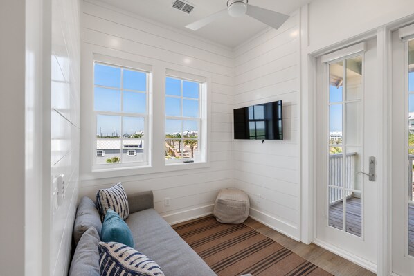 Una habitación con pared blanca, ventilador de techo, ventana grande, televisor de pantalla plana, sofá con cojines y puerta que da a una terraza exterior.