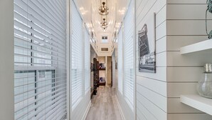 A narrow hallway with white blinds, a light wooden floor, and a ceiling with a chandelier. There are shelves on the right wall and a door at the end of the hallway.