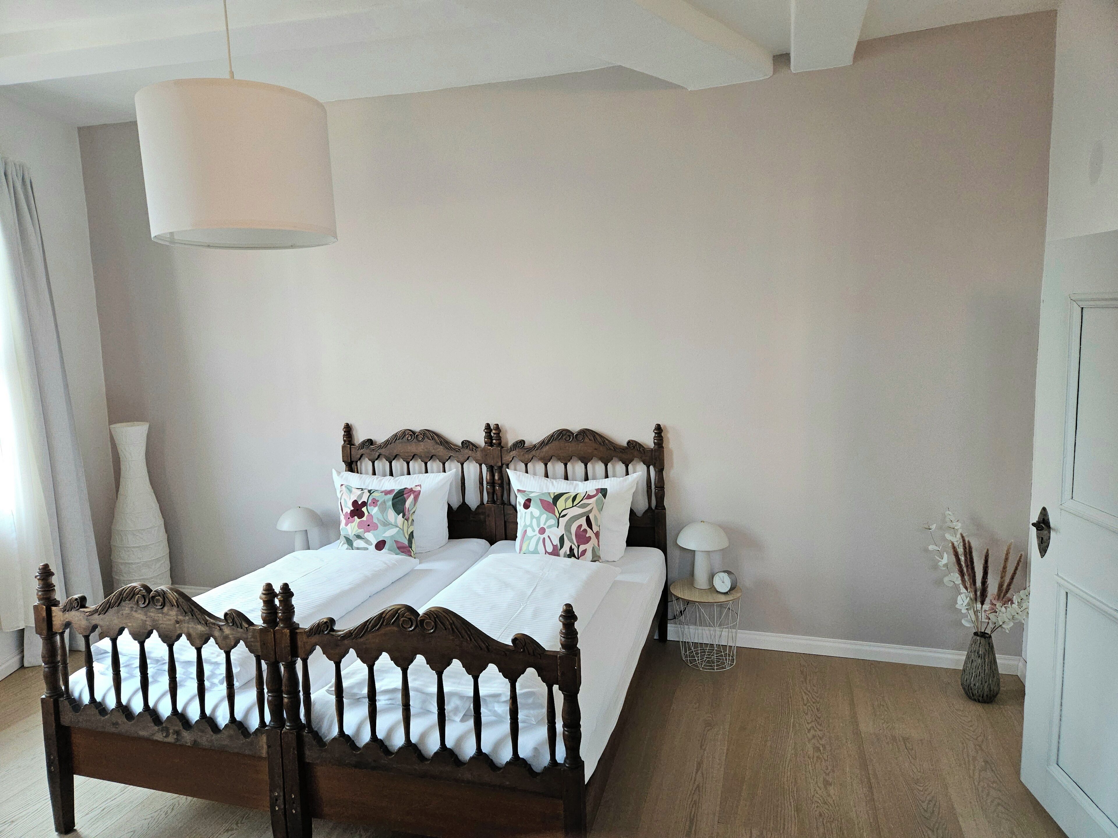 A bedroom with a wooden bed, white bedding, a bedside table with a lamp, and a vase with dried plants.