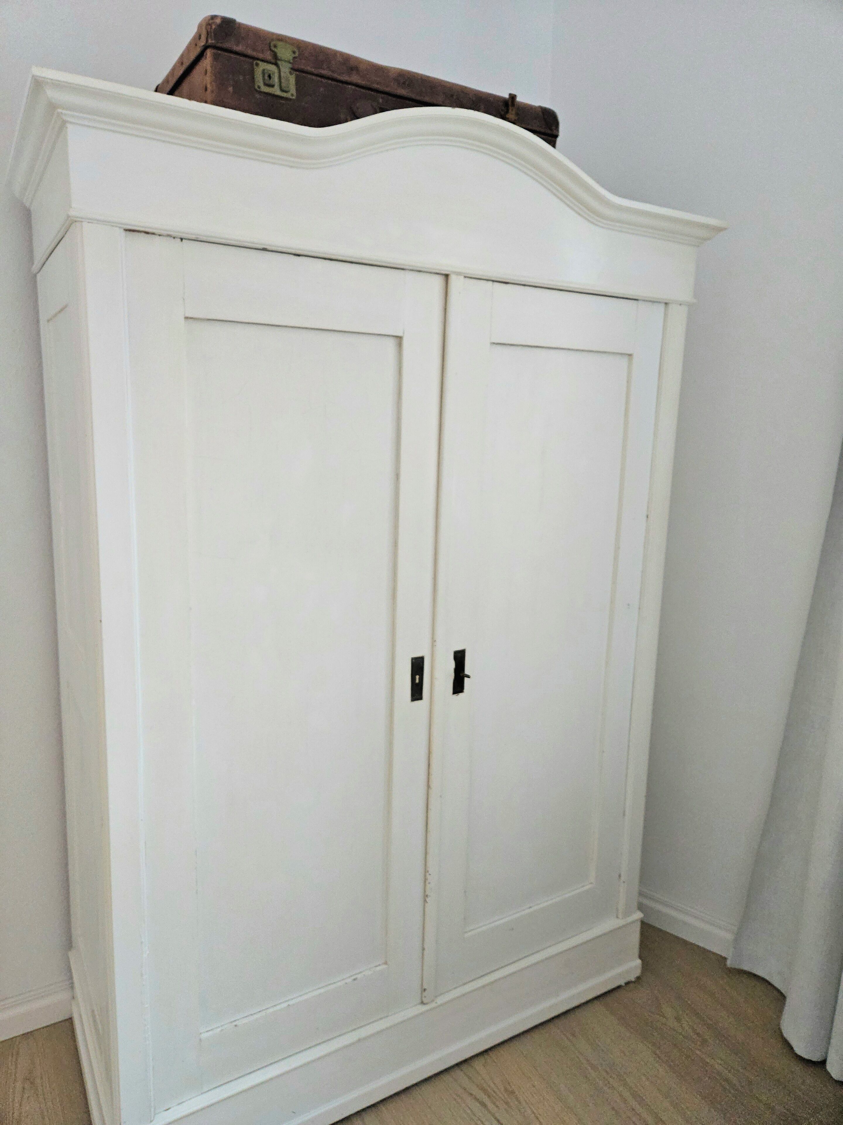 Une armoire blanche à deux portes, surmontée d’un petit coffre.