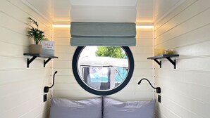 A bedroom with a round window, a bed with two pillows, a bedside table with a plant, and a shelf with a sign.