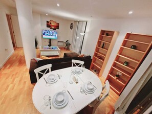A dining area with a round table set for four, a television mounted on the wall, and a bookshelf to the right.