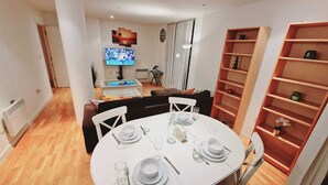 A dining area with a round table set for four, a television mounted on the wall, and a bookshelf to the right.