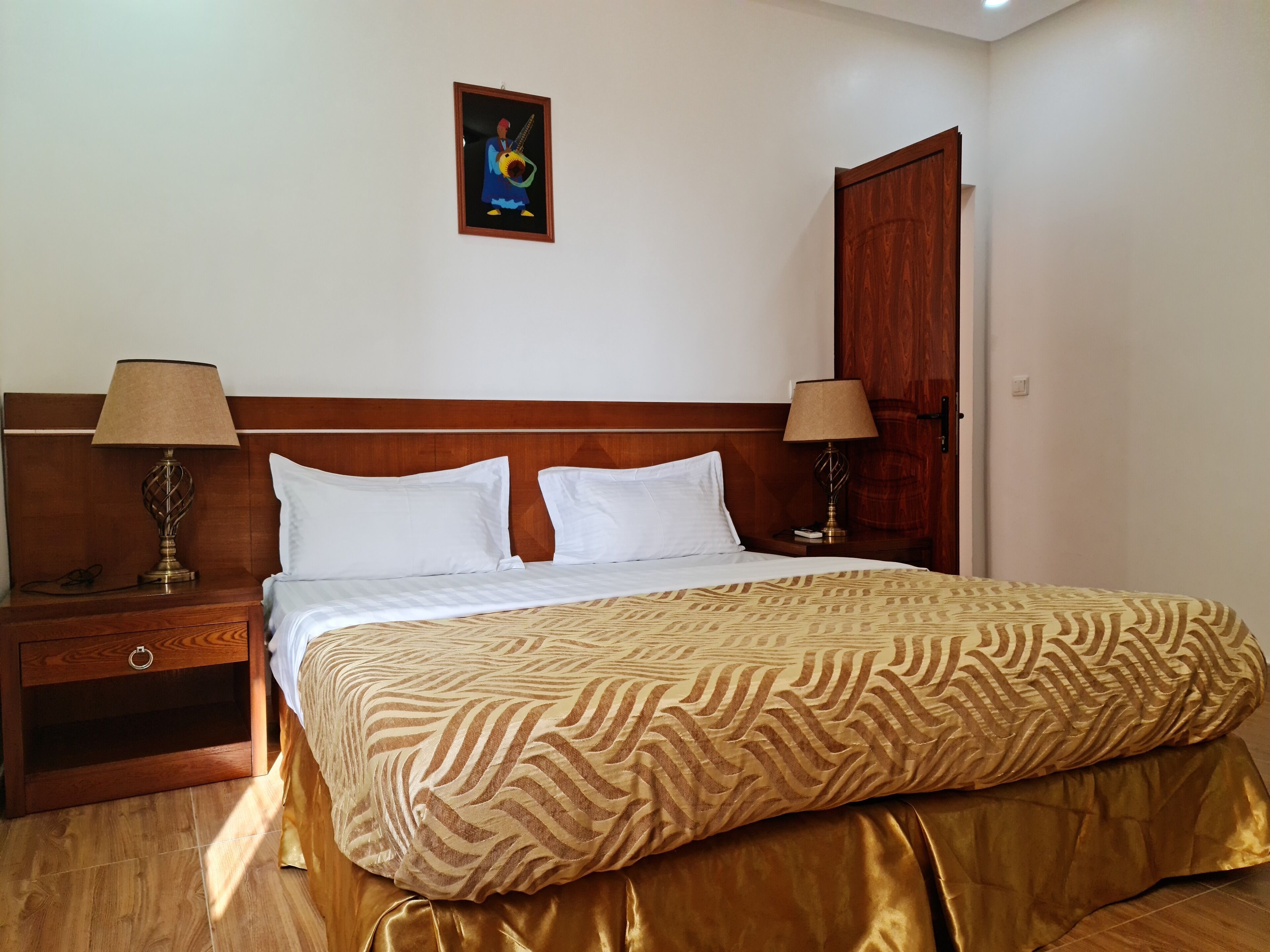 Una habitación de hotel con cabecera de madera, dos mesitas de noche con lámparas, una cama con colcha estampada y un cuadro en la pared.