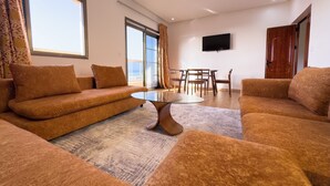 A spacious living room with a large sofa set, a glass coffee table, and a dining area with chairs and a table. - Oasis Seaside by Barok (Dakar)