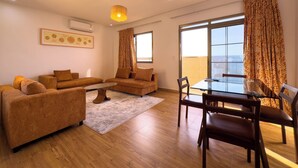 A modern living room with a sofa set, a glass dining table with chairs, a patterned rug, and a large window with curtains. - Oasis Seaside by Barok (Dakar)