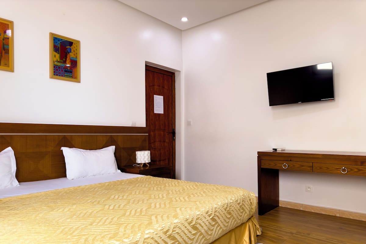 A hotel room with a bed, a wooden headboard, a TV mounted on the wall, a wooden dresser, and a door with a wooden panel.