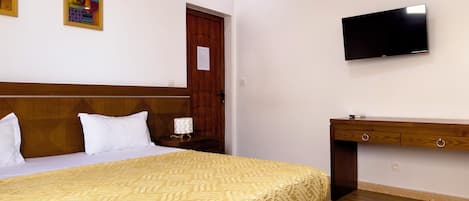Habitación de hotel con cama, cabecera de madera, televisor montado en la pared, cómoda de madera y una puerta con panel de madera.