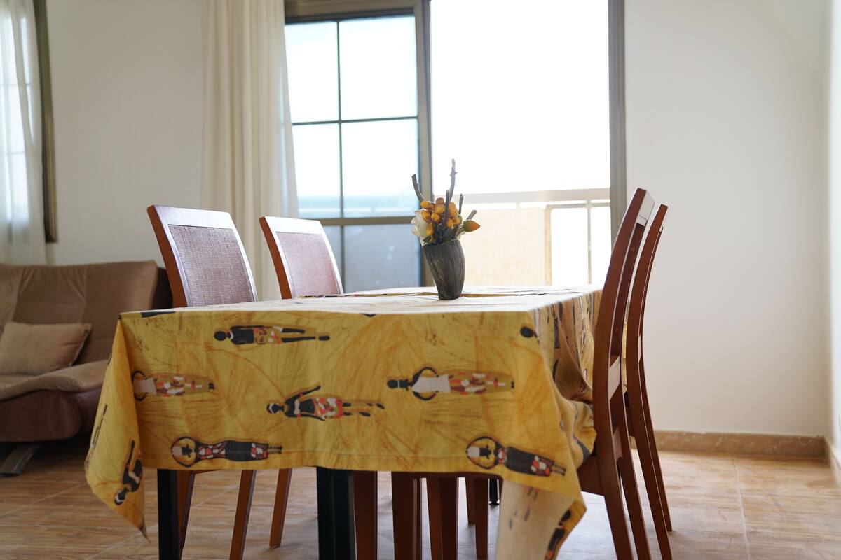 A dining table with a yellow tablecloth, surrounded by wooden chairs, in a room with a large window and a sofa.