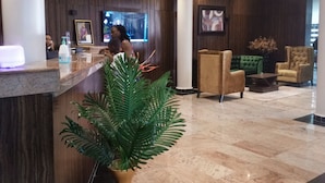 Zona de recepción con suelo de mármol, mostrador de recepción, una planta grande en maceta y un área de estar con sillones y una mesa de centro.