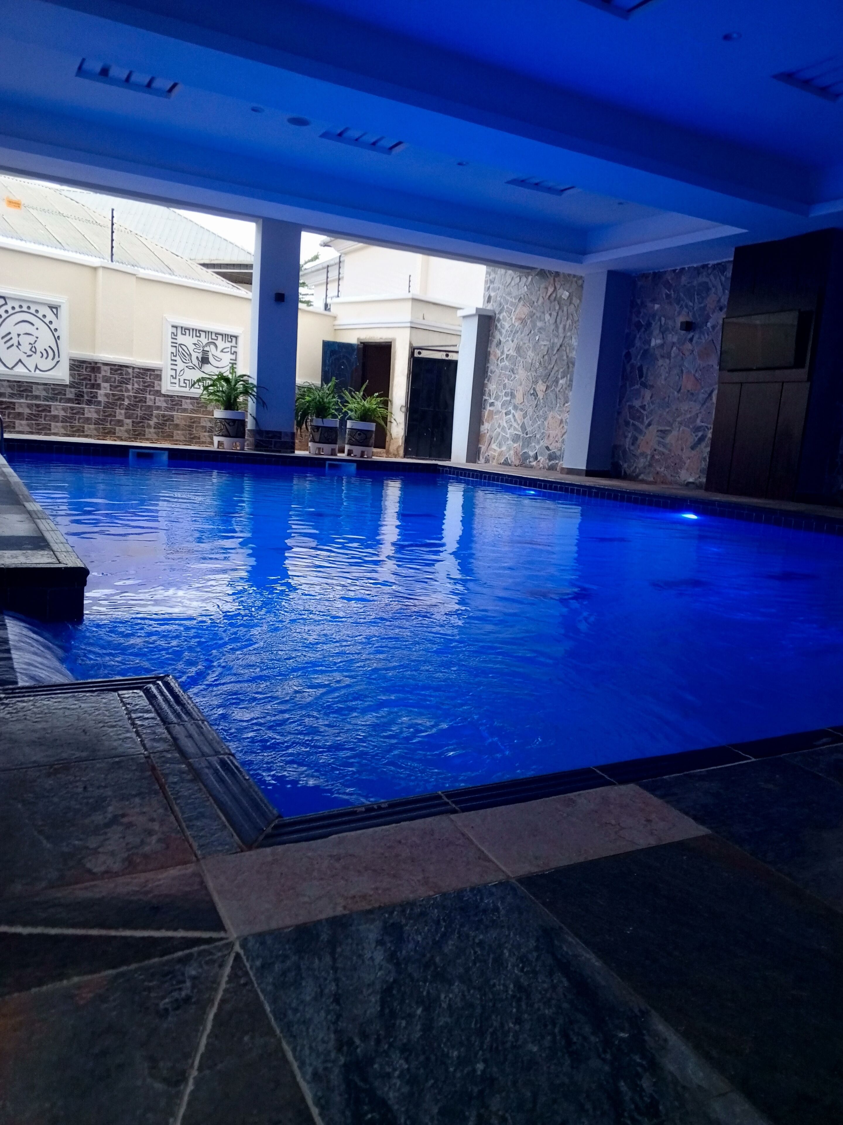 Una piscina interior con un elemento acuático azul, paredes de piedra y plantas en macetas.