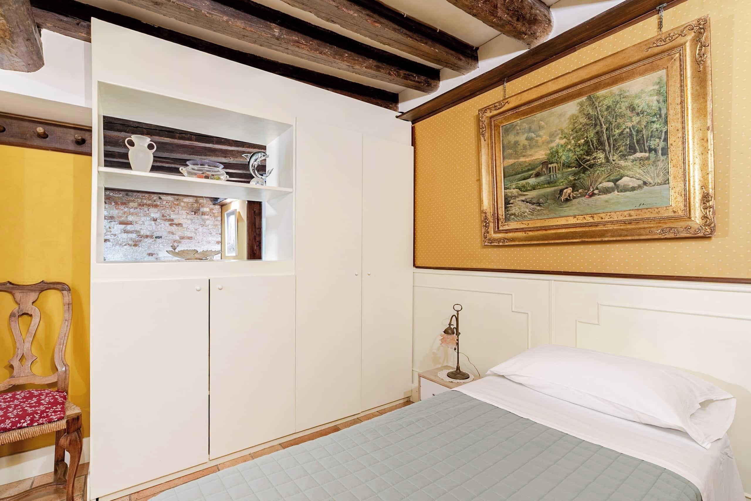 A bedroom with a bed, a wooden chair, a shelf with decorative items, a painting, and a brick wall.