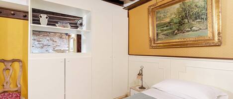A bedroom with a bed, a wooden chair, a shelf with decorative items, a painting, and a brick wall.
