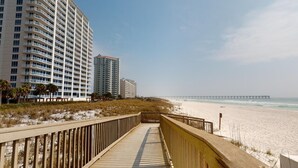 Een houten looppad dat naar een strand leidt, met hoogbouw op de achtergrond.