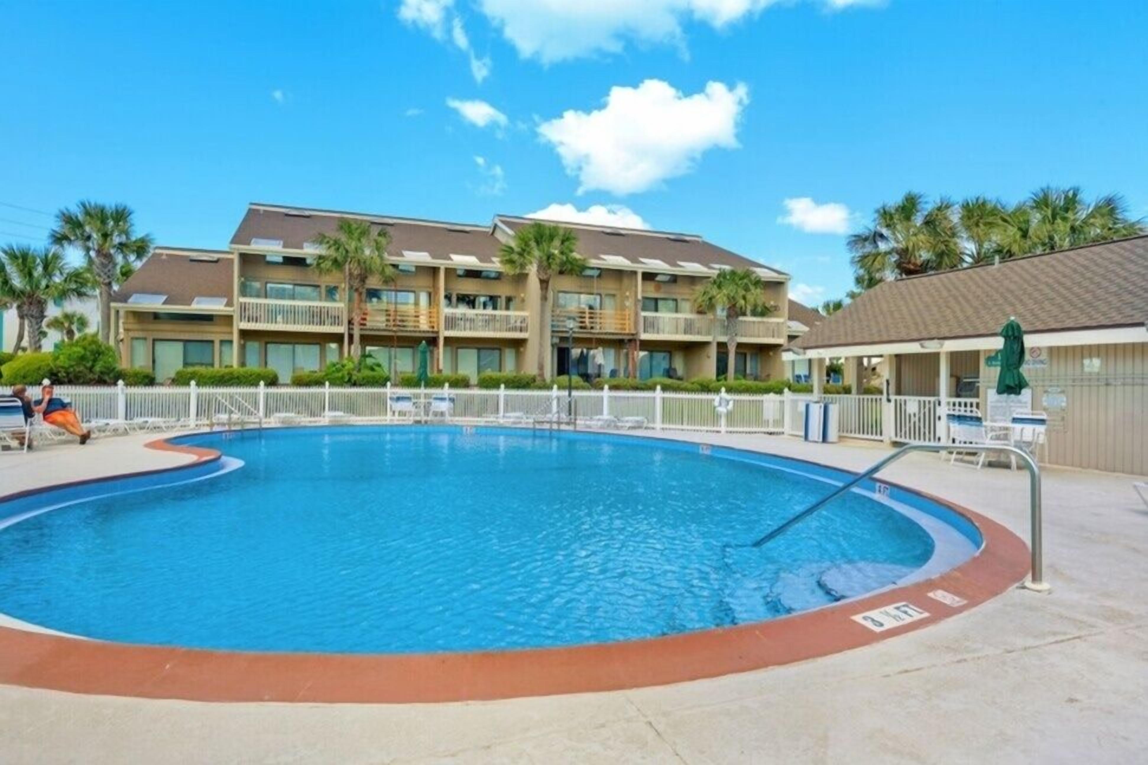 A swimming pool with a white fence, palm trees, and a building with balconies in the background.
