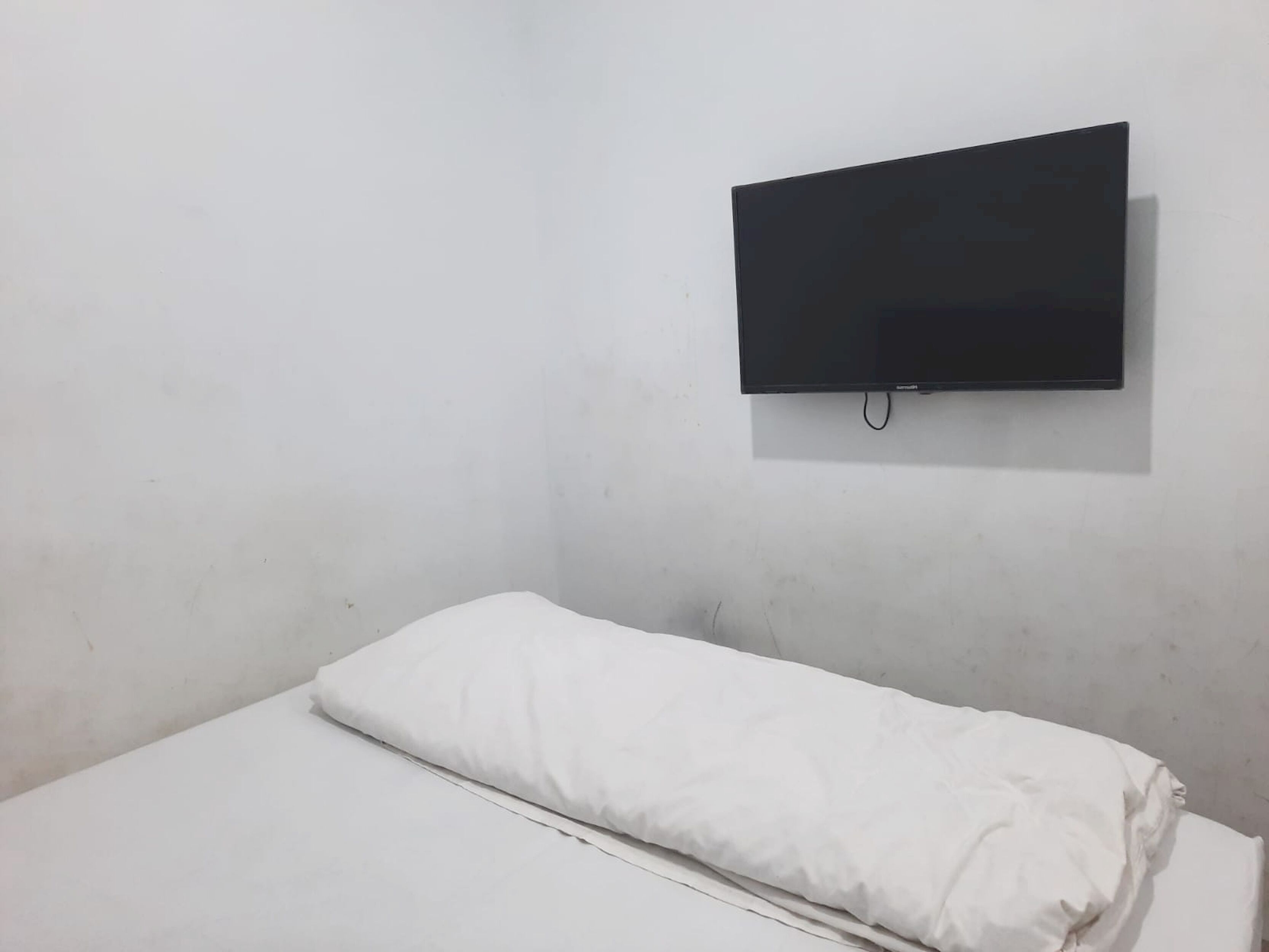 A single bed with white bedding in a room with a plain white wall and a mounted television.