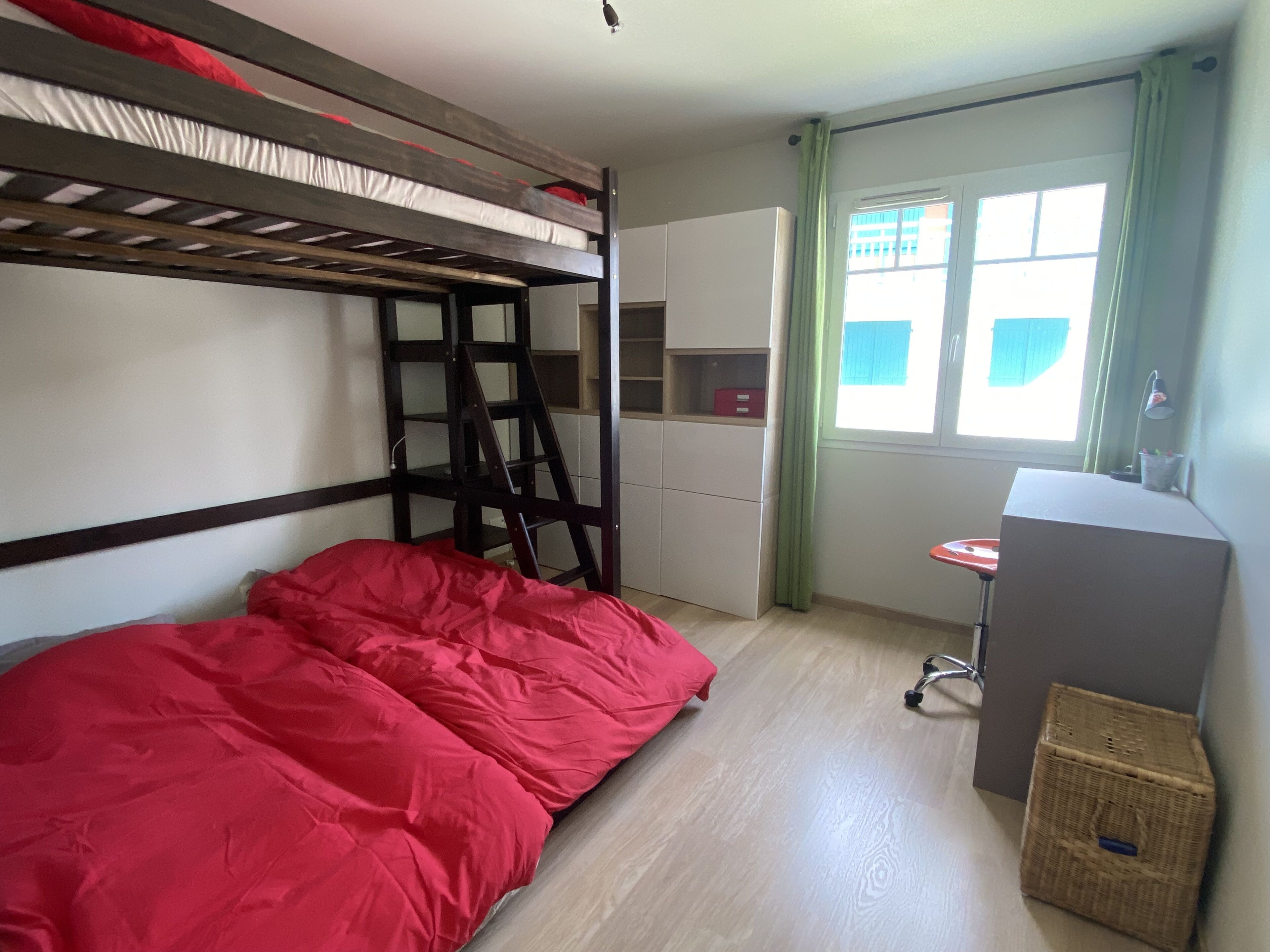 Una cama litera con ropa de cama roja, un escritorio con una silla, una cesta de mimbre y una ventana con cortinas.