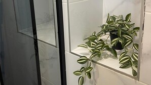 A bathroom with white marble tiles, a glass shower enclosure, and a potted plant on a shelf.