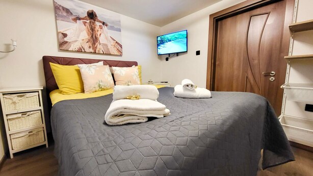 A neatly made bed with a grey quilt, yellow and floral pillows, and two folded towels.