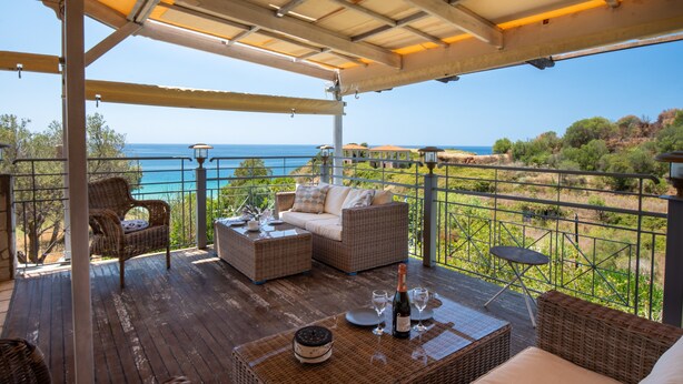 Un espacio de estar al aire libre cubierto con muebles de mimbre, una mesa con copas de vino y vistas al mar.