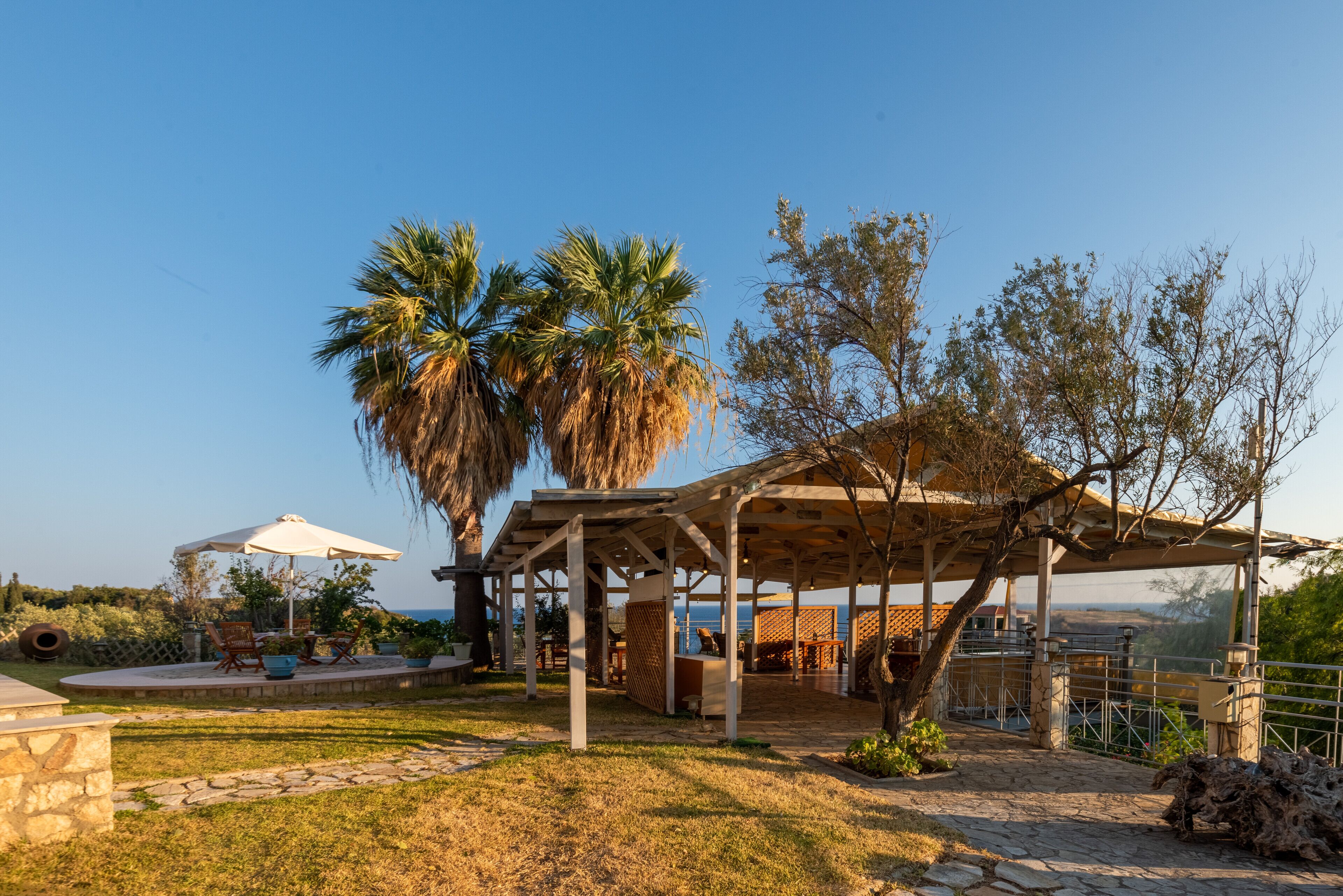 A covered outdoor seating area with stone flooring, surrounded by palm trees and a view of the sea.