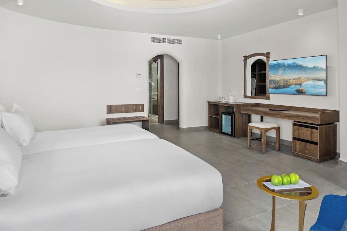 Une chambre d’hôtel moderne avec un grand lit, un bureau avec une chaise, une petite table avec des pommes et un tableau au mur.