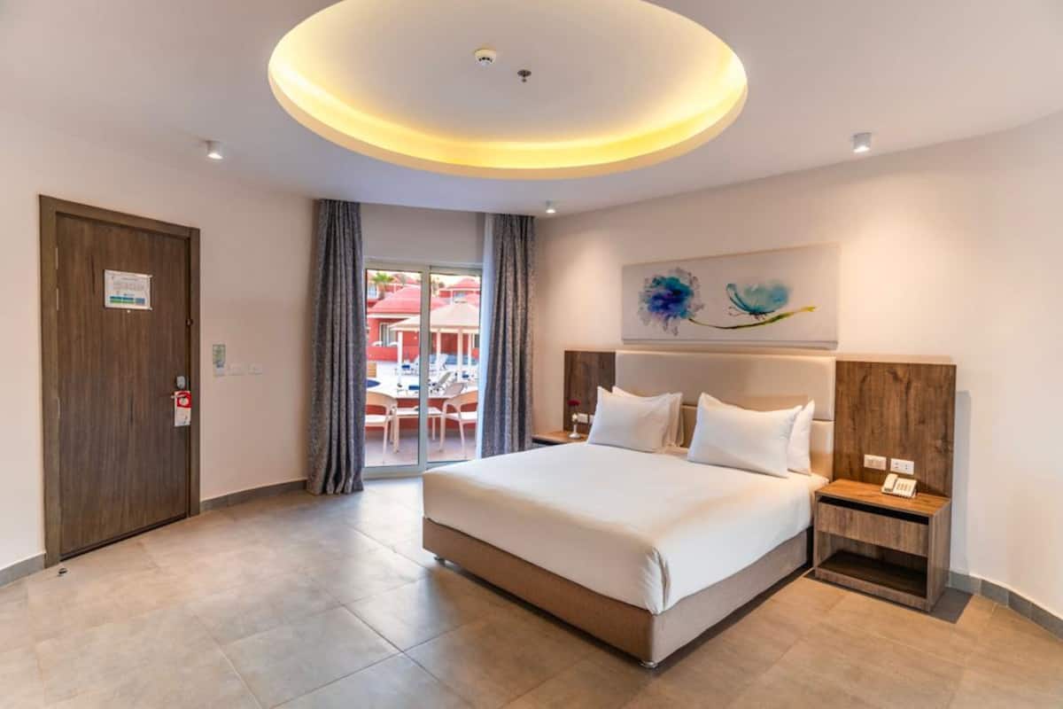 Une chambre d’hôtel avec un grand lit, une tête de lit en bois, une table de chevet avec un téléphone et une vue sur l’extérieur grâce à une porte vitrée.