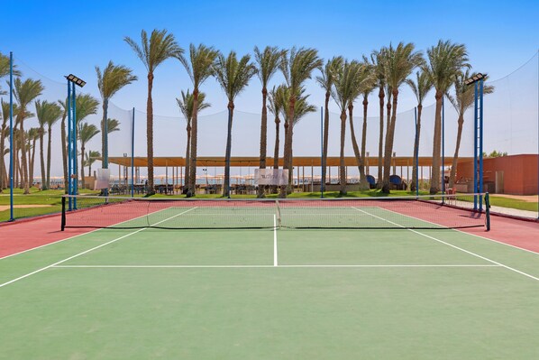 A tennis court with a net, surrounded by palm trees and a building in the background.