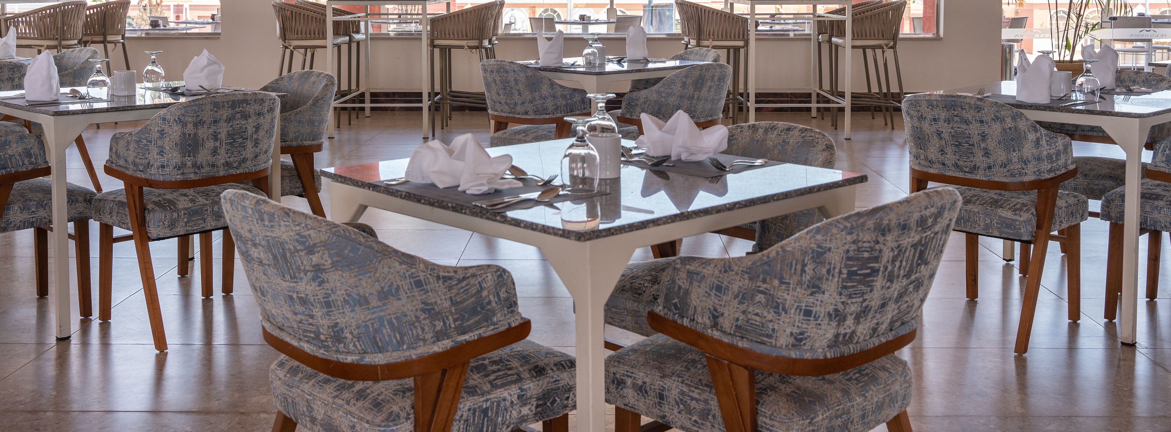 Uma sala de jantar com mesas redondas e cadeiras azuis estampadas, disposta em um espaço amplo com luz natural proveniente de grandes janelas.