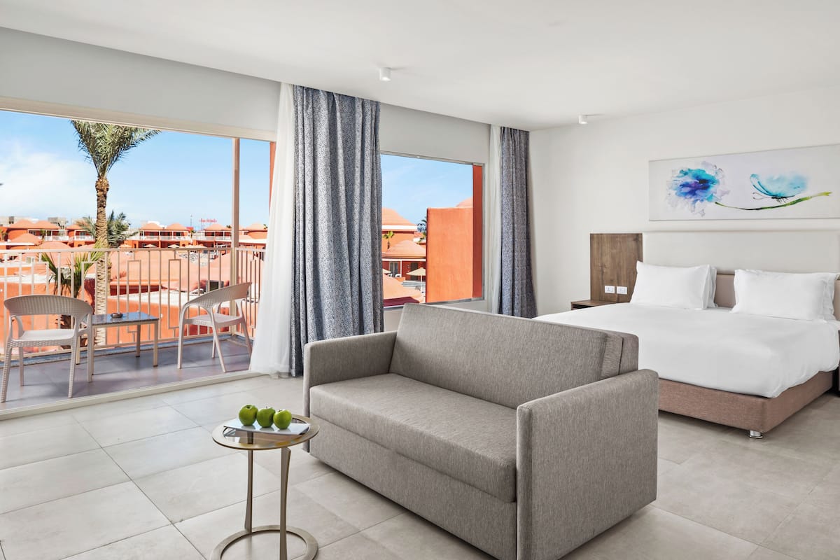 Une chambre d’hôtel avec un lit, un canapé, une petite table avec des pommes, et un balcon avec des chaises et un palmier.