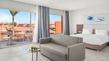 Una habitación de hotel con cama, sofá, una mesita con manzanas y un balcón con sillas y una palmera.