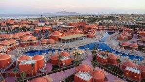 Un complexe hôtelier comprenant plusieurs bâtiments à toits rouges, une piscine centrale et offrant une vue sur la mer et les montagnes en arrière-plan.