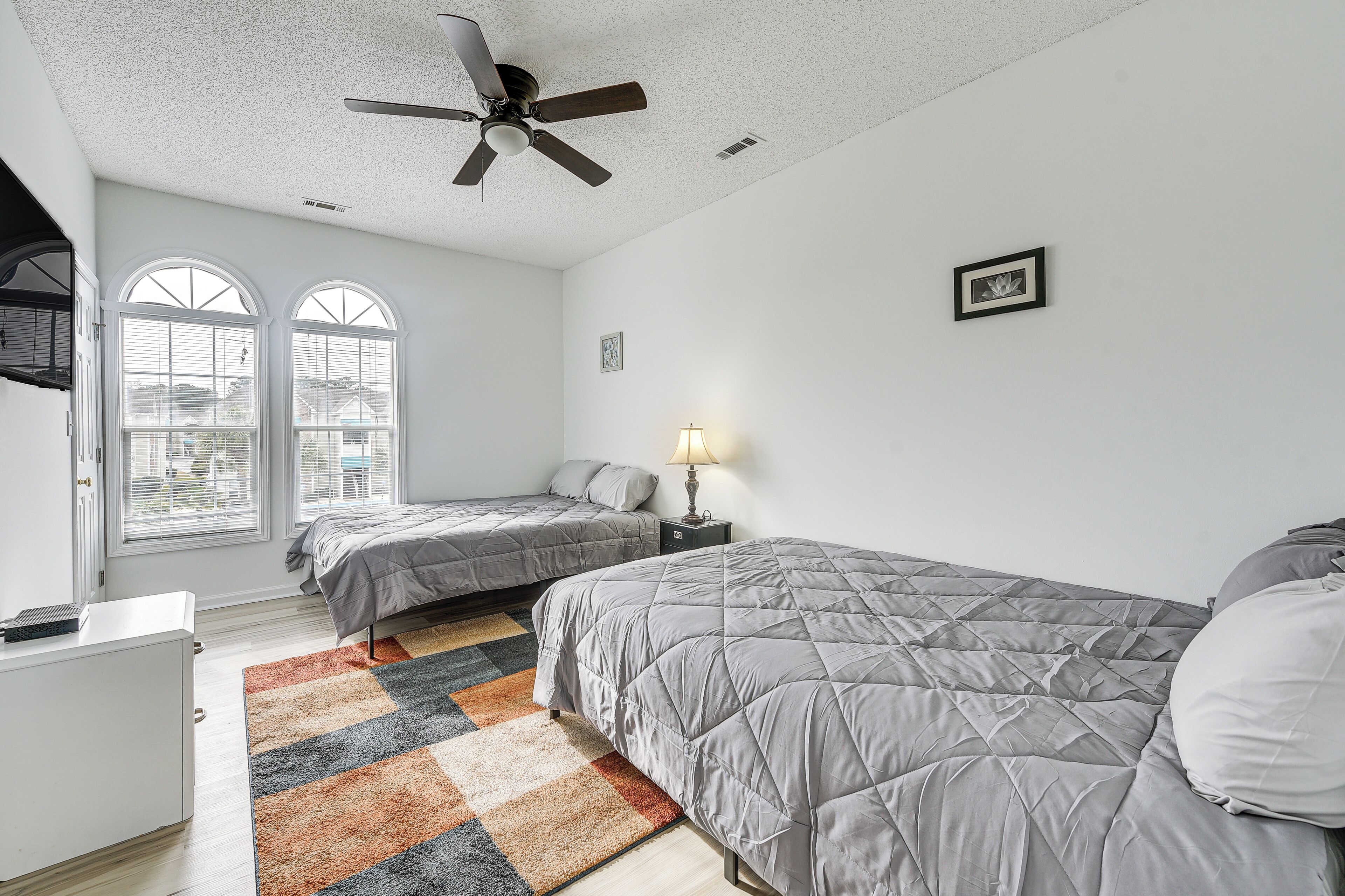 A bedroom with two beds, a ceiling fan, a window with blinds, a lamp, and a rug.