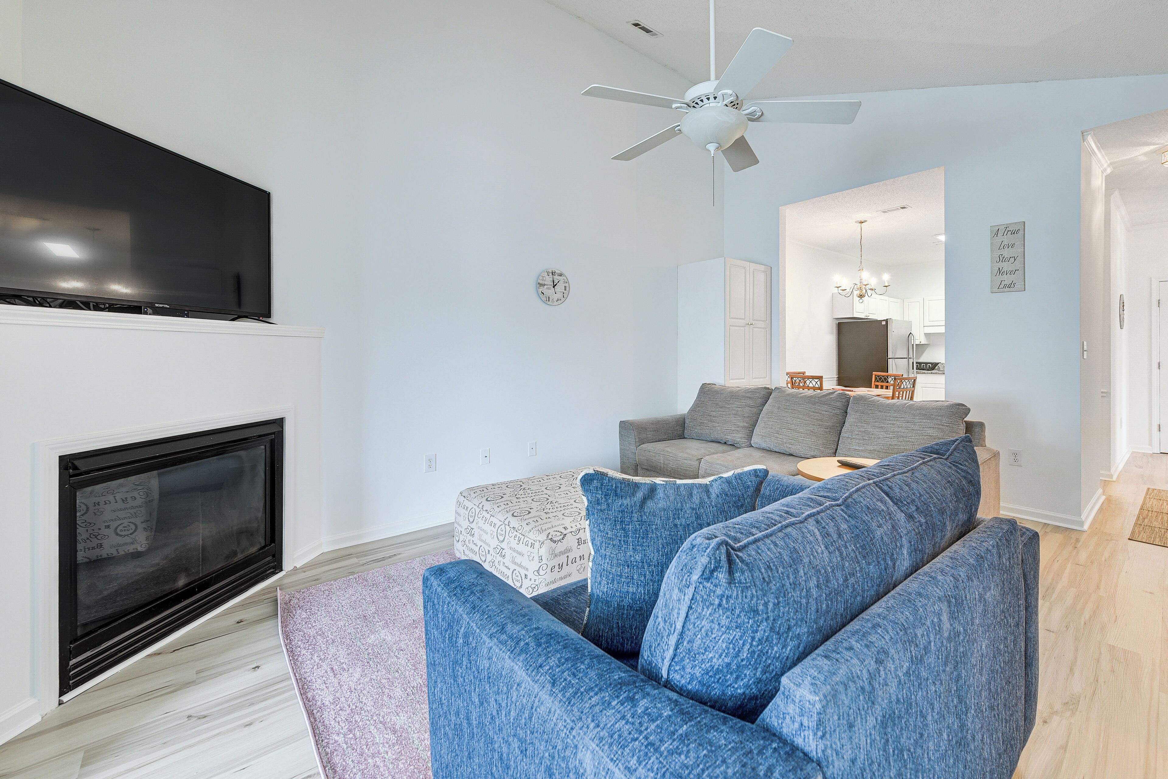 A modern living room with a flat-screen TV, a ceiling fan, a fireplace, and a seating area with a grey sofa and blue cushions.