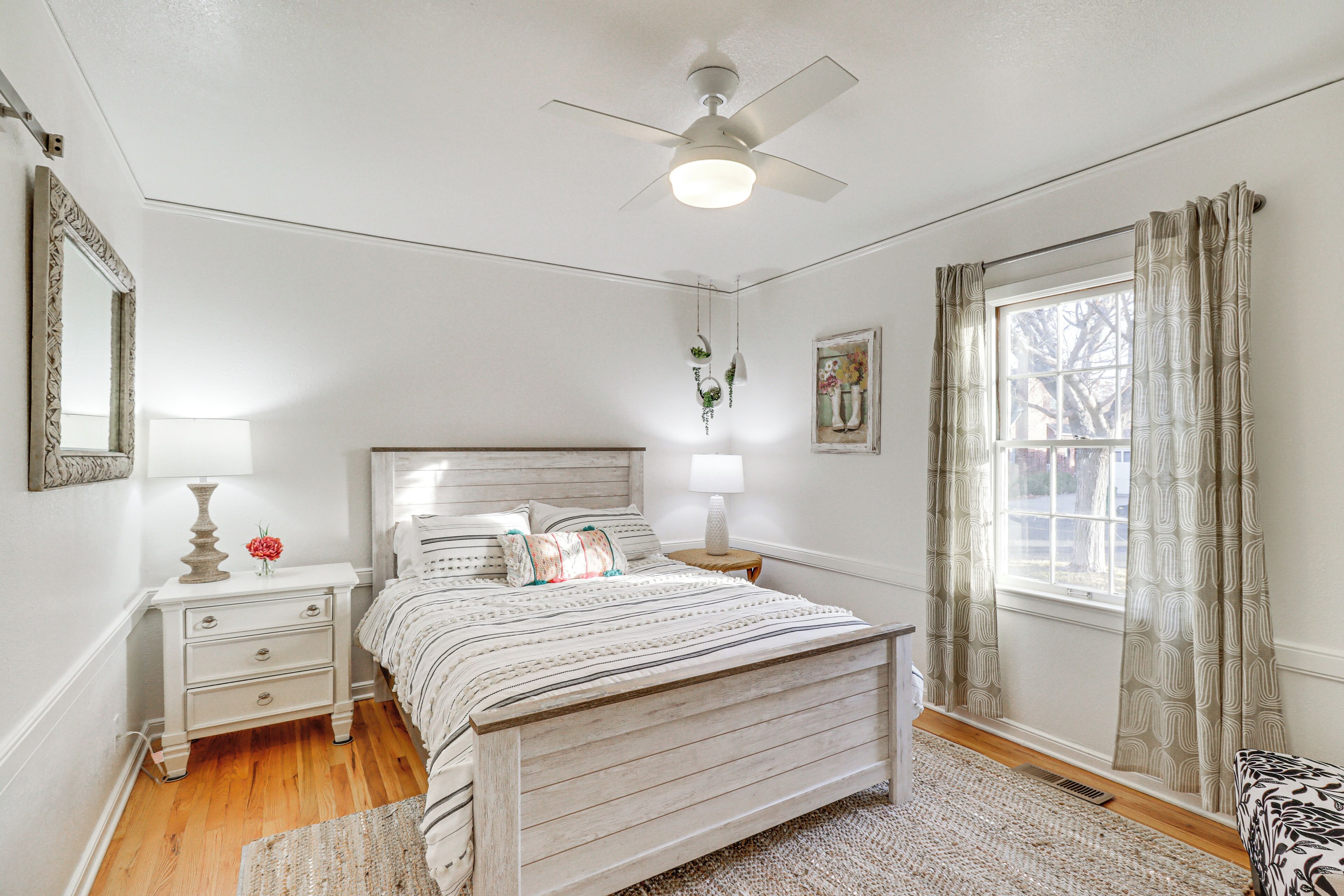 Une chambre à coucher avec un lit, des tables de chevet, un ventilateur de plafond, une fenêtre avec des rideaux et un tableau encadré au mur.