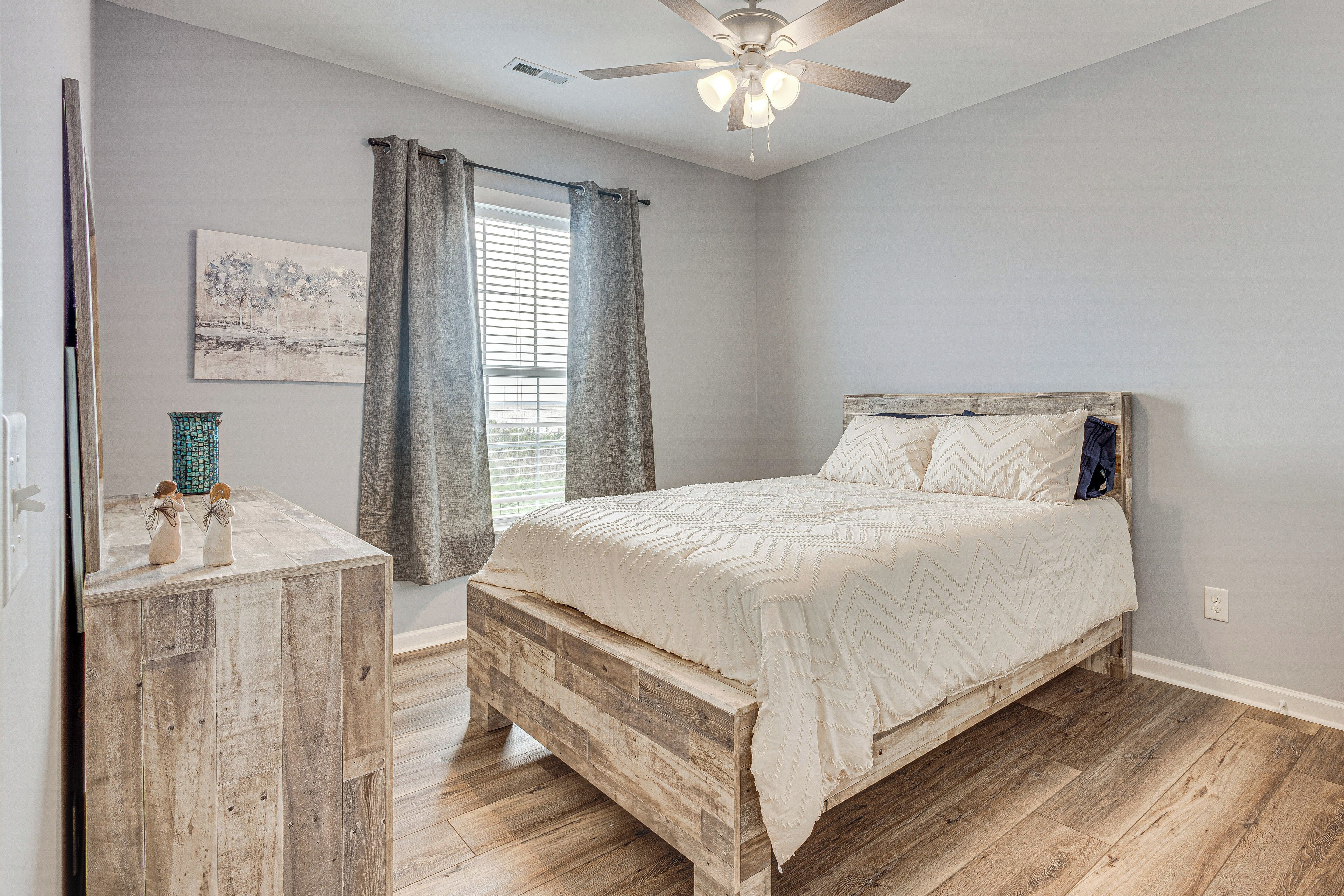 Quarto com cama de madeira, ventoinha no teto, janela com cortinas e pequena mesa com objetos decorativos.