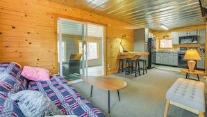 A cozy cabin interior with a wooden floor, a sofa, a table, and a kitchen area. - Oscoda Cabin w/ Charcoal Grill: Walk to Lake Huron (Oscoda)
