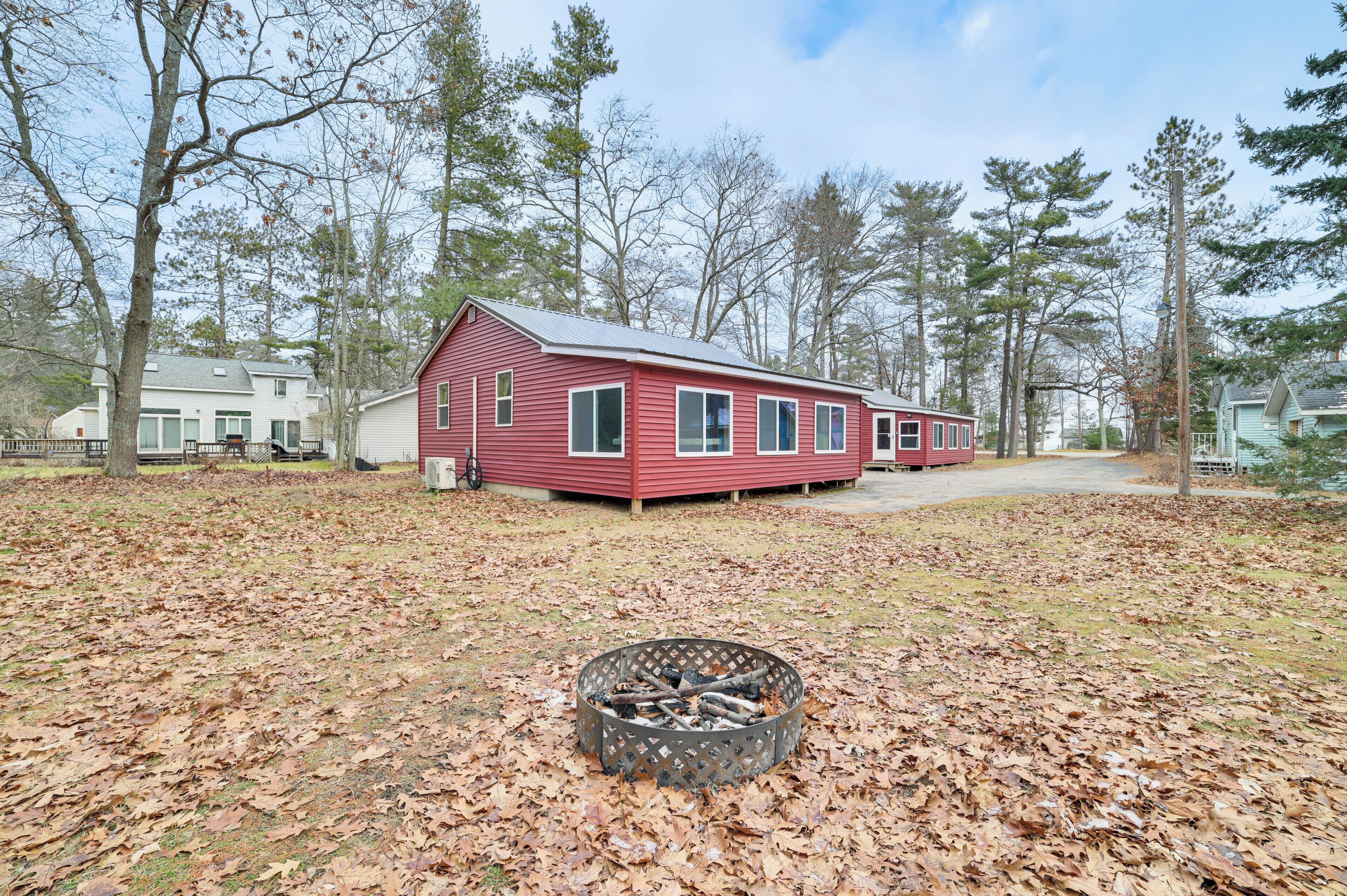 A red cabin with a fire pit in the yard, surrounded by trees and a white house in the background.