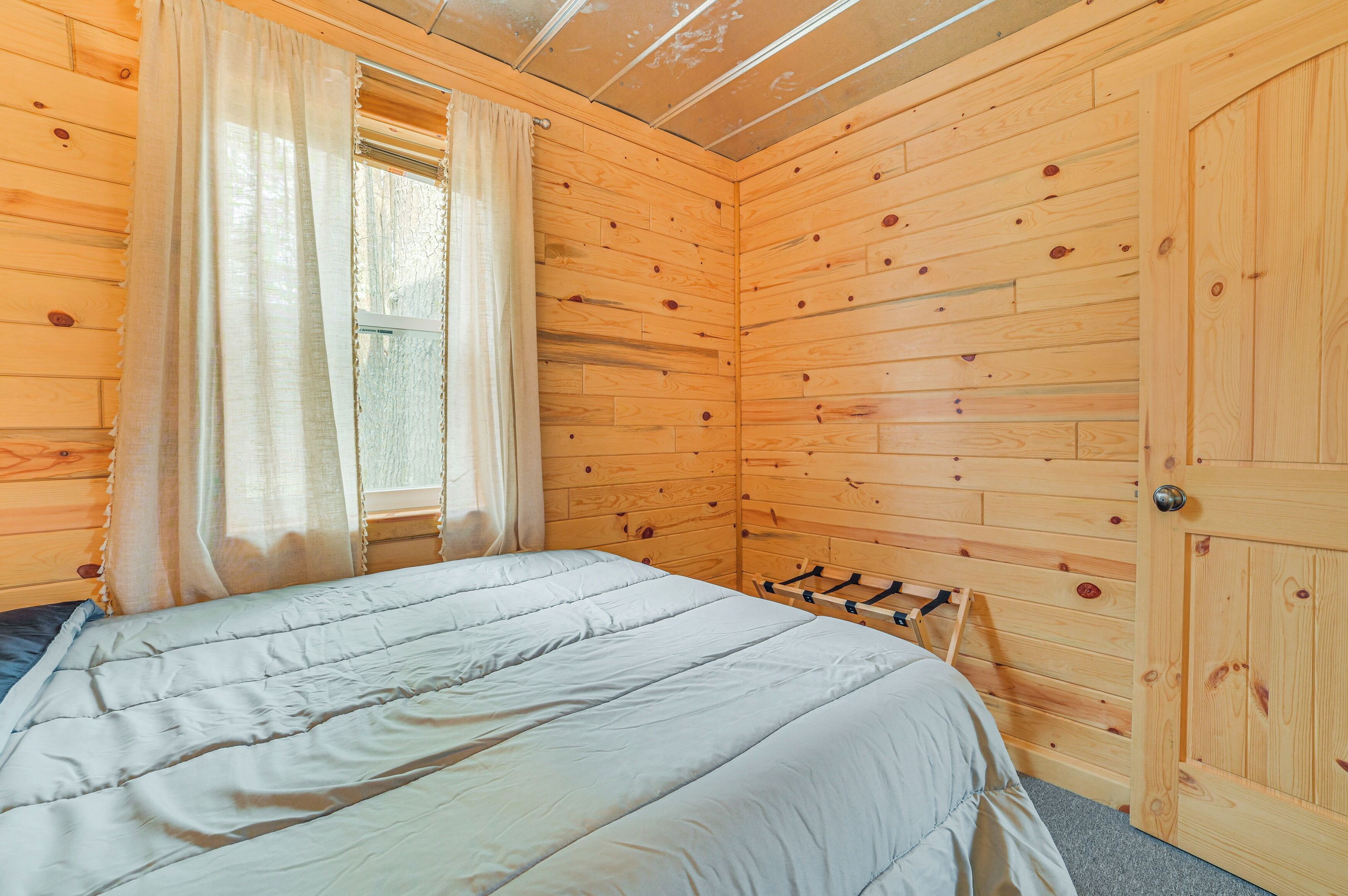 A wooden cabin room with a bed, a window with curtains, and a door.