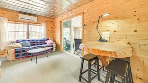 A cozy room with a wooden interior, a sofa, a table, and a bar stool. - Oscoda Cabin w/ Charcoal Grill: Walk to Lake Huron (Oscoda)
