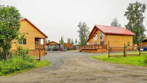 A rustic wooden cabin with a dirt road leading to it, surrounded by greenery and other cabins. - Soldotna Fishing Cabin ~ Half-mi to Kenai River! (Soldotna)