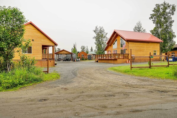 A rustic wooden cabin with a dirt road leading to it, surrounded by greenery and other cabins. - Soldotna Fishing Cabin ~ Half-mi to Kenai River! (Soldotna)