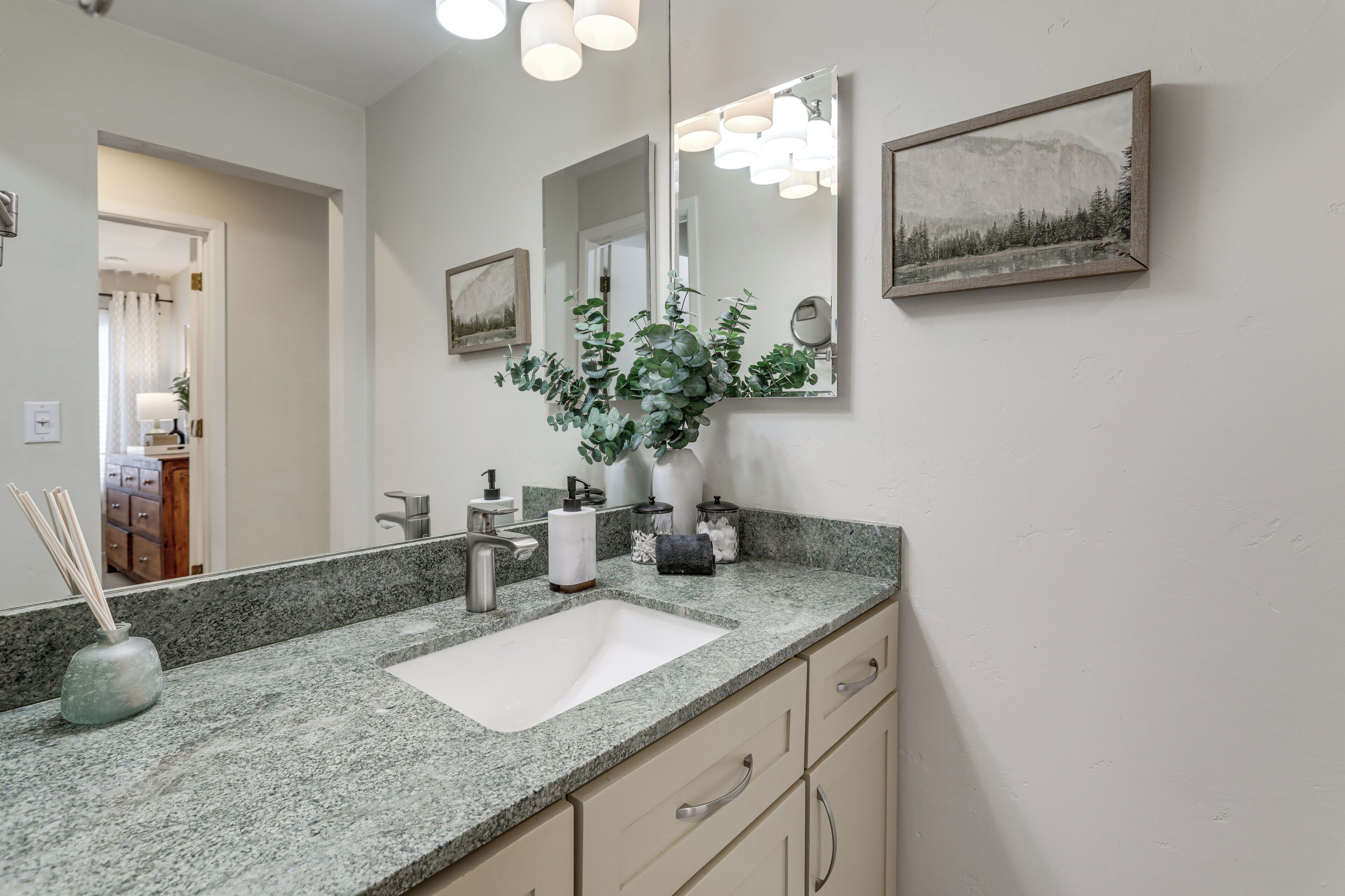 A bathroom with a granite countertop, a white sink, a mirror, and a framed landscape painting on the wall.