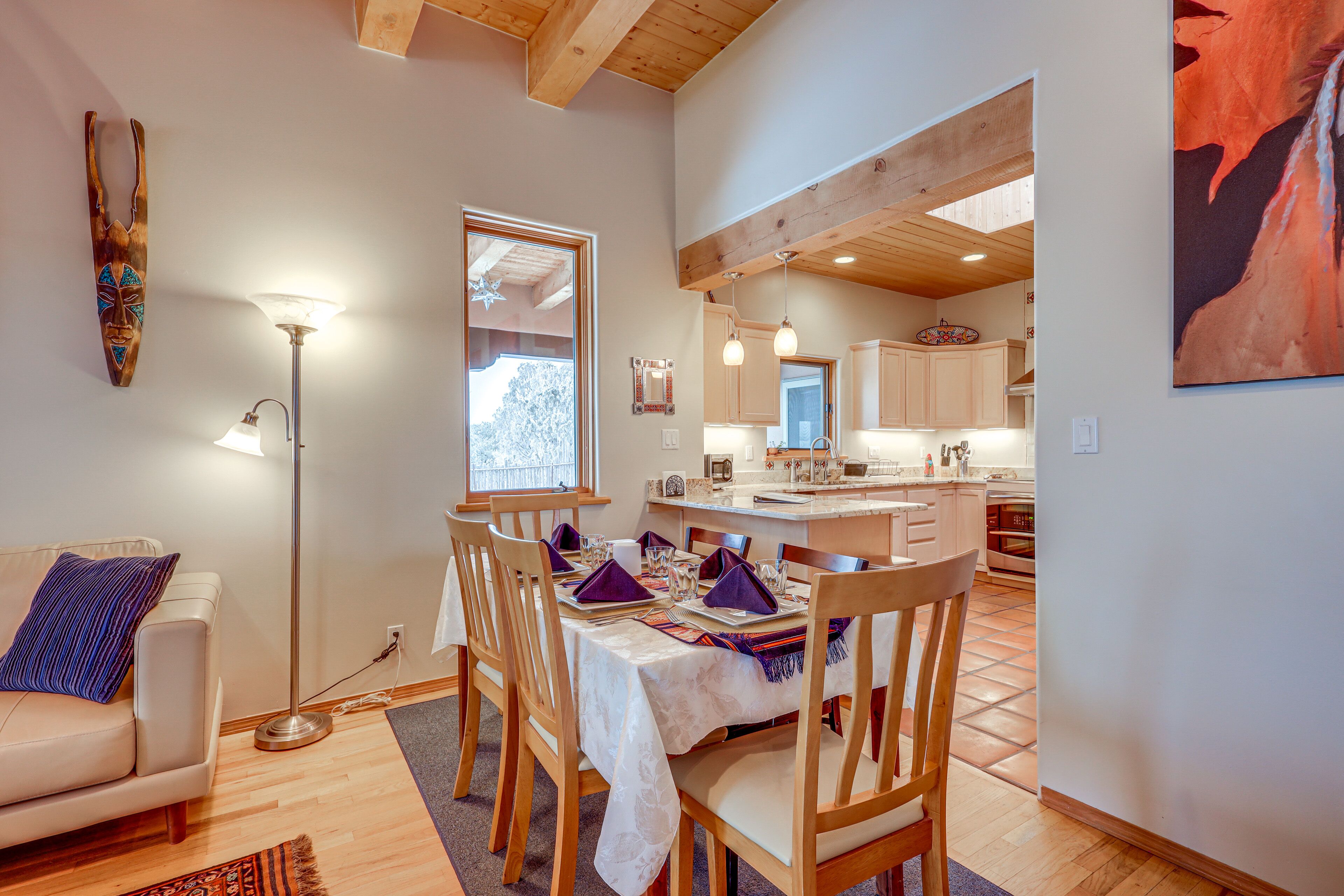 A dining area with a table set for four, wooden chairs, a floor lamp, and a view into the kitchen.