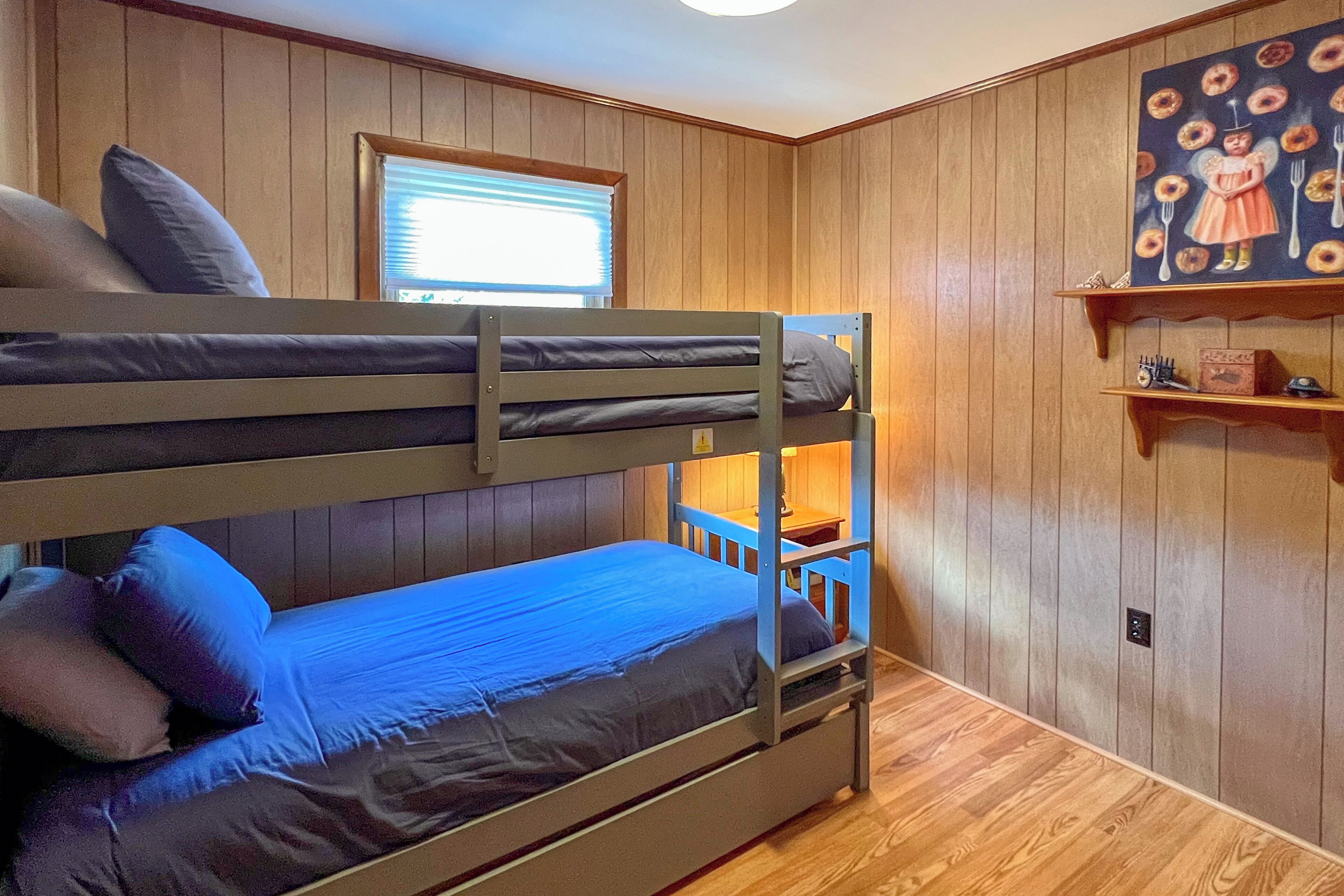 A bunk bed room with wooden walls, a window with blinds, and a shelf with a painting.