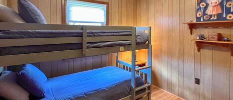 A bunk bed room with wooden walls, a window with blinds, and a shelf with a painting.