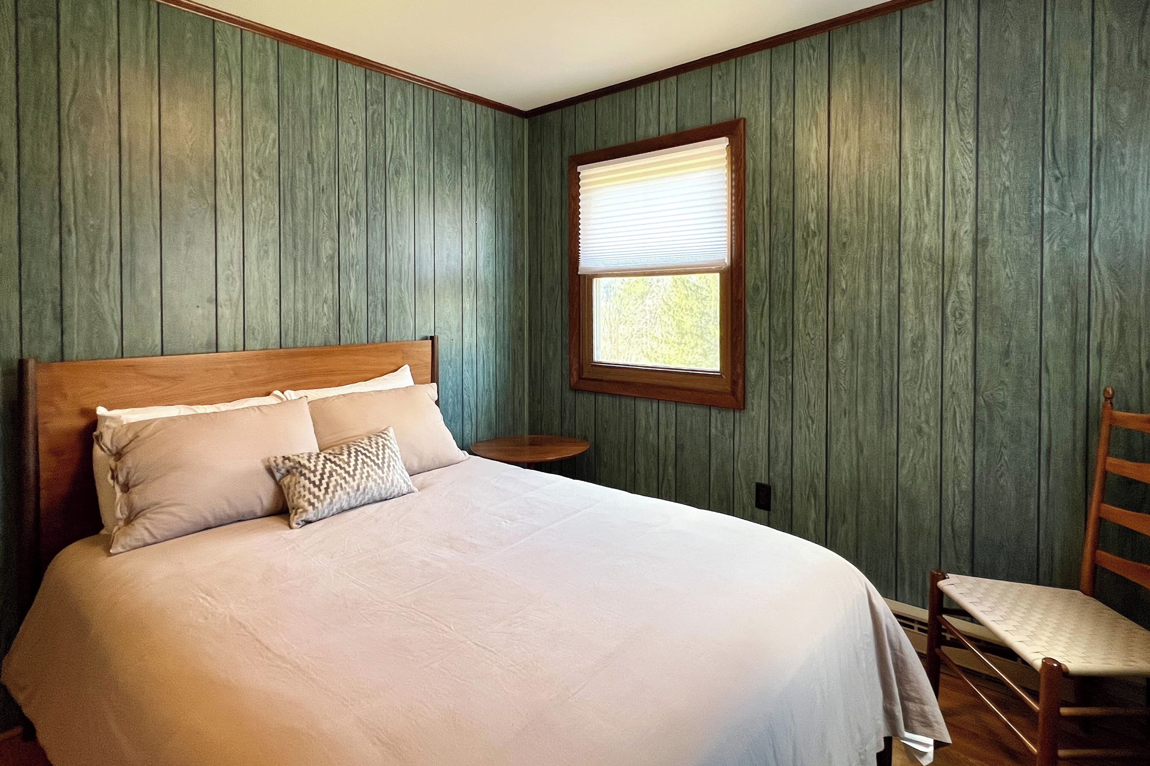 A neatly made bed with a wooden headboard, a small round table beside it, and a window with blinds.