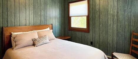 A neatly made bed with a wooden headboard, a small round table beside it, and a window with blinds.